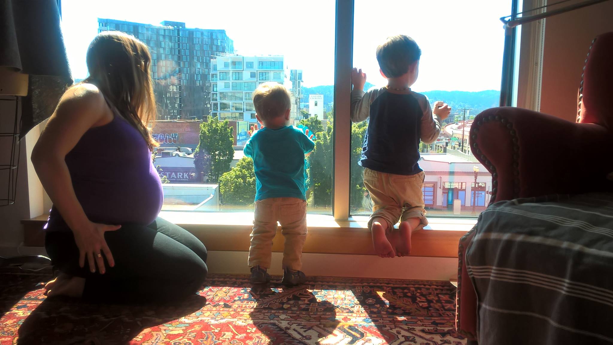 Nicole heavily pregnant crouching beside two small boys looking out a high rise window over Portland Oregon