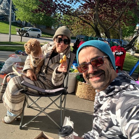 Nicole and Adam sitting in camp chairs at their garage sale with Poppy the Cavapoo a box of donuts and coffee — selling everything before leaving for Europe.
