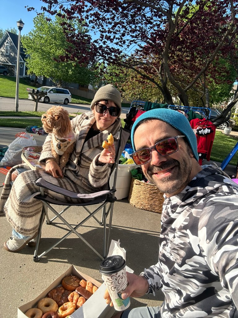 Nicole and Adam sitting in camp chairs at their garage sale with Poppy the Cavapoo a box of donuts and coffee — selling everything before leaving for Europe.