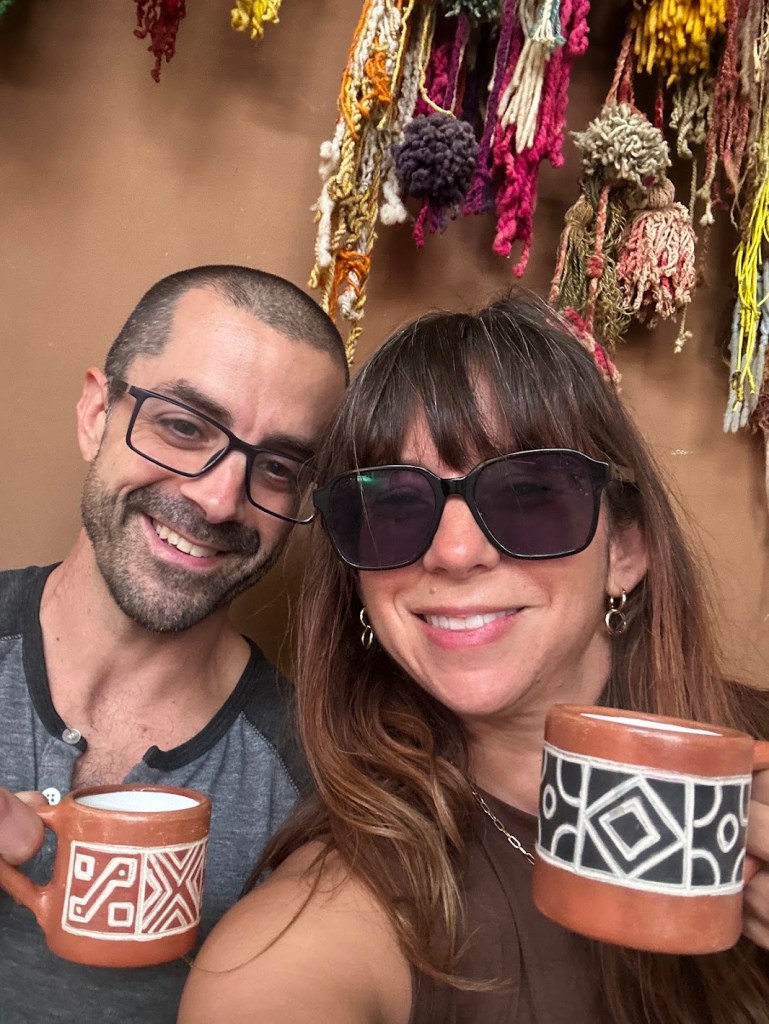Nicole and Adam holding matching artisan ceramic mugs and smiling — captured somewhere on their travels together. The couple behind Adventures We Seek who sold their house packed their kids and their Cavapoo Poppy and are 55 days away from heading to Europe with no return date set.