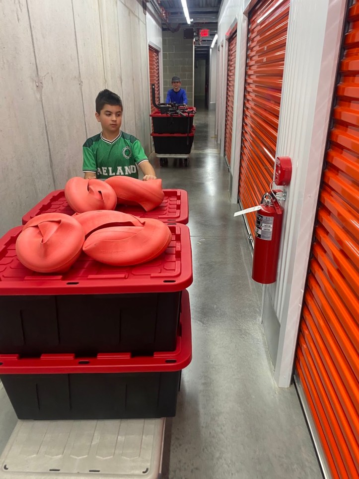 Jake wearing an Ireland jersey and Gavin pushing bins through a storage unit hallway... two kids helping their family store their belongings before selling their home and traveling to Europe.