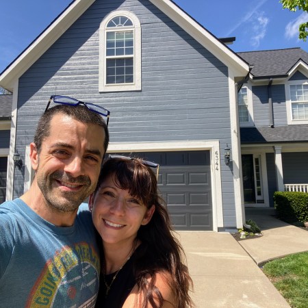 Nicole and Adam smiling in front of their beautiful blue Kansas home the day it went on the market... coming soon on Zillow and MLS before they sell everything and travel to Europe with their family.