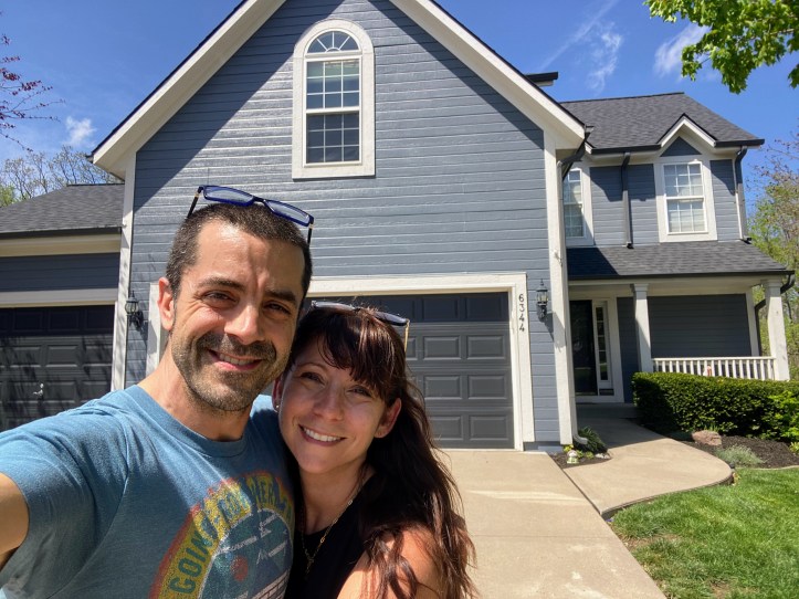Nicole and Adam smiling in front of their beautiful blue Kansas home the day it went on the market... coming soon on Zillow and MLS before they sell everything and travel to Europe with their family.
