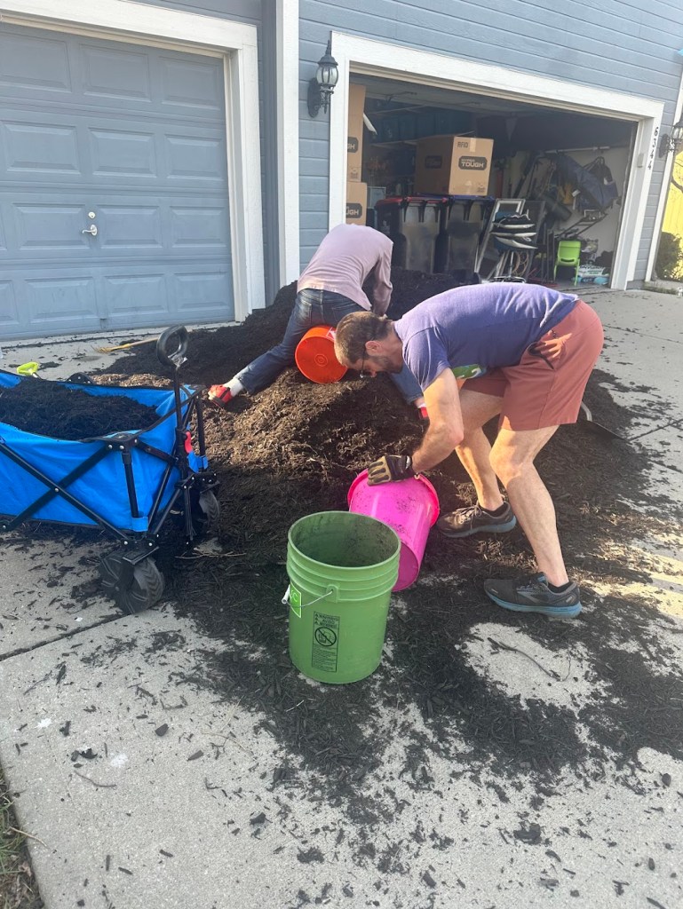 Adam and Gavin shoveling six yards of mulch from the driveway into colorful buckets to spread throughout the yard — 53 days before the family leaves for Europe.