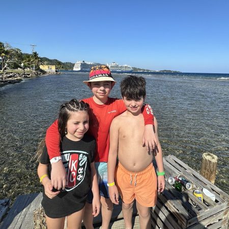 Avilene, Gavin and Jake smiling together on a dock with cruise ships and the ocean behind them — three kids who were born for adventure.
