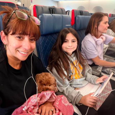 Mom holding a small curly brown dog wrapped in a red blanket on a Delta flight, sitting next to her young daughter who is wearing earbuds and holding a tablet, flying over the North Atlantic on the way to Europe for worldschooling