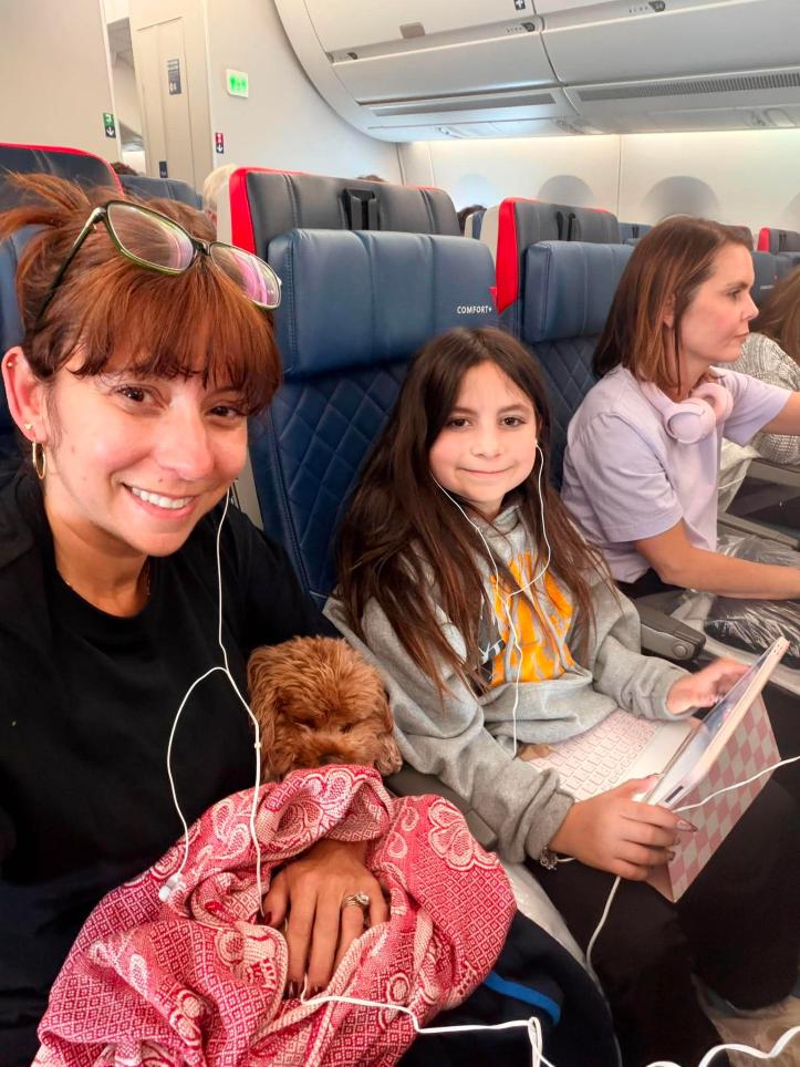 Mom holding a small curly brown dog wrapped in a red blanket on a Delta flight, sitting next to her young daughter who is wearing earbuds and holding a tablet, flying over the North Atlantic on the way to Europe for worldschooling