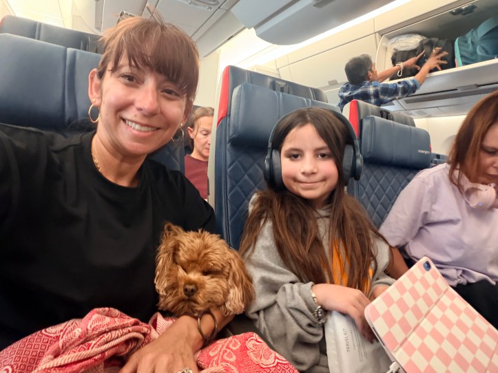 Nicole and her daughter Avilene smile for a selfie on a Delta flight with a small golden Cavapoo tucked under a blanket between them.