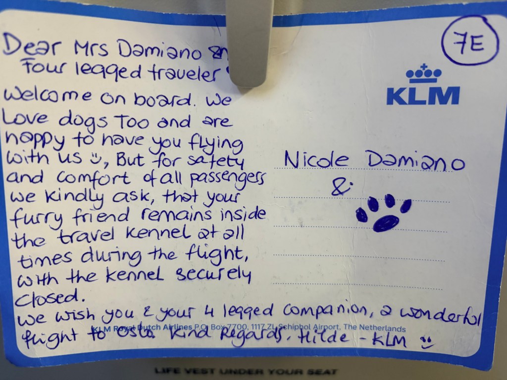 A handwritten KLM notecard addressed to Nicole Damiano welcoming her and her dog on board, kindly asking that the dog remain in the travel kennel during the flight, and wishing them a wonderful flight to Oslo. Signed by Hilde from KLM with a hand drawn paw print.