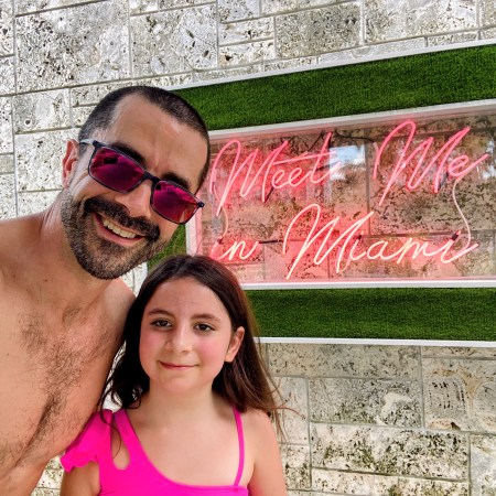 Adam and Avilene smiling in front of a pink neon Meet Me in Miami sign — a dad and his daughter soaking up the last big adventure before the biggest adventure of their lives.