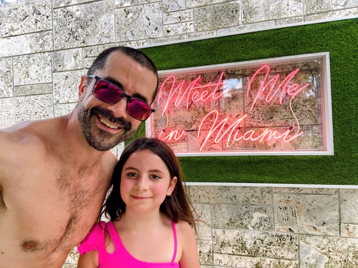 Adam and Avilene smiling in front of a pink neon Meet Me in Miami sign — a dad and his daughter soaking up the last big adventure before the biggest adventure of their lives.