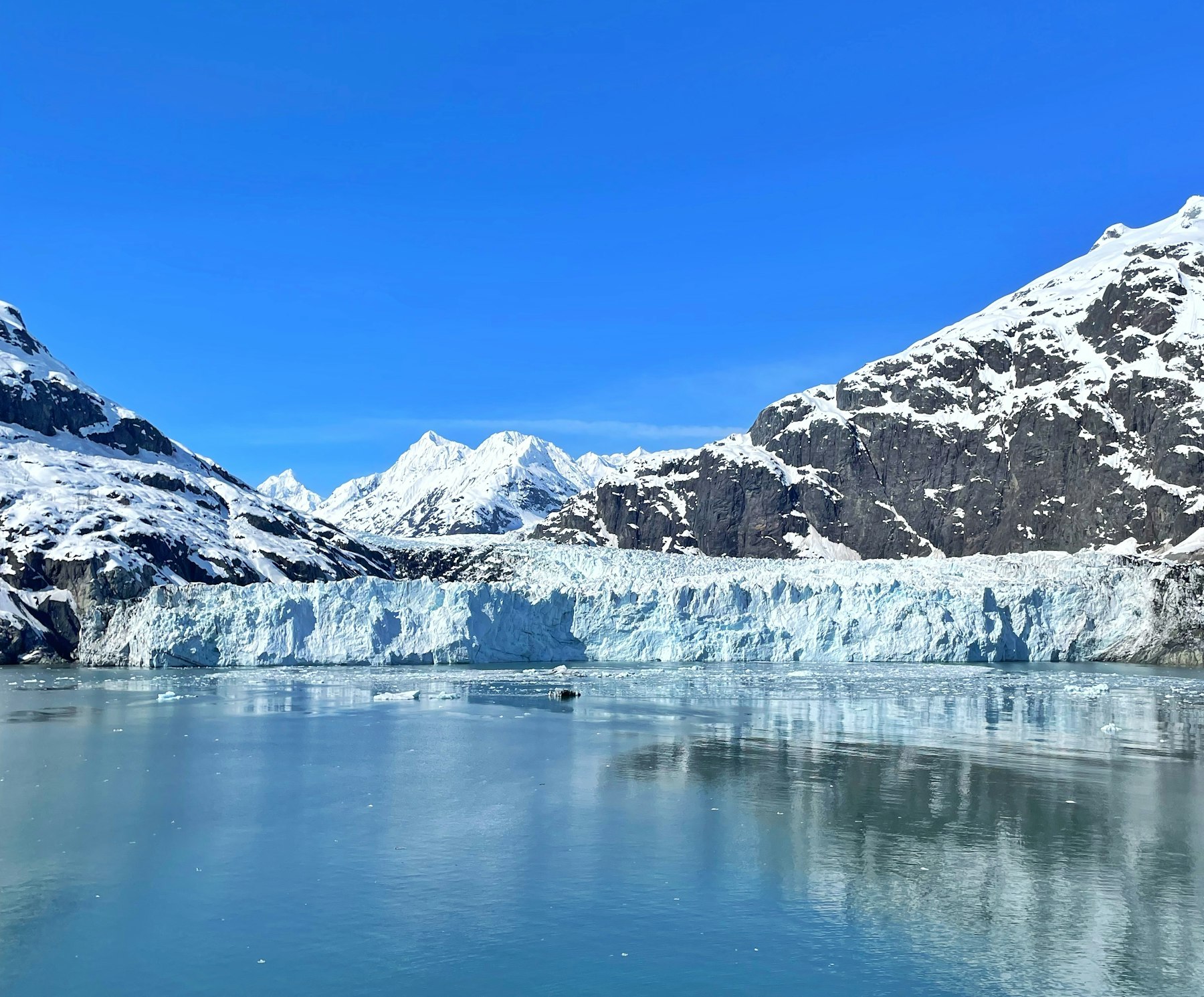 A wide glacier front meeting calm water below snow covered peaks in Glacier Bay Alaska