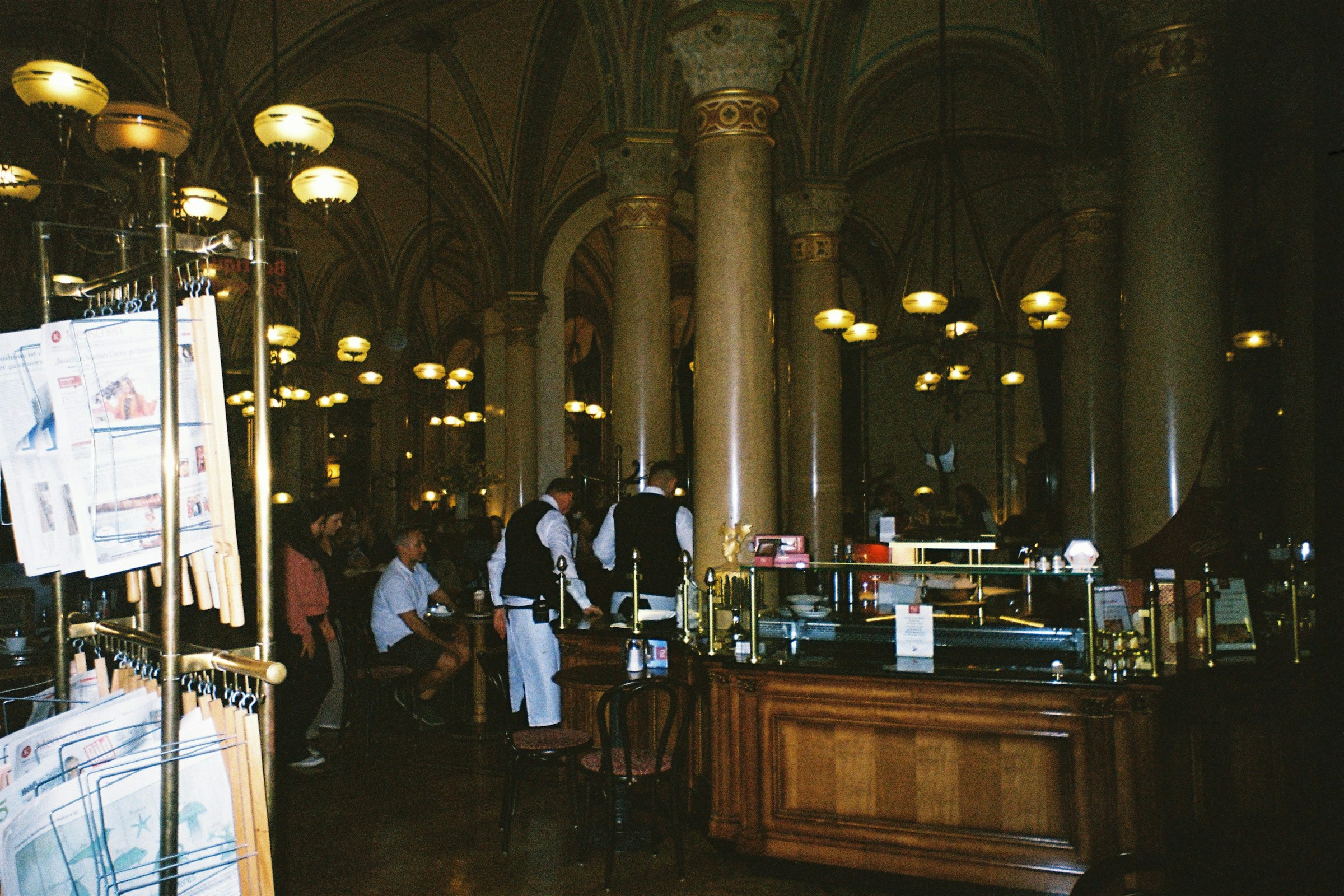 The vaulted interior of Cafe Central in Vienna with marble columns, globe lamps, waiters in waistcoats and newspapers on a rack