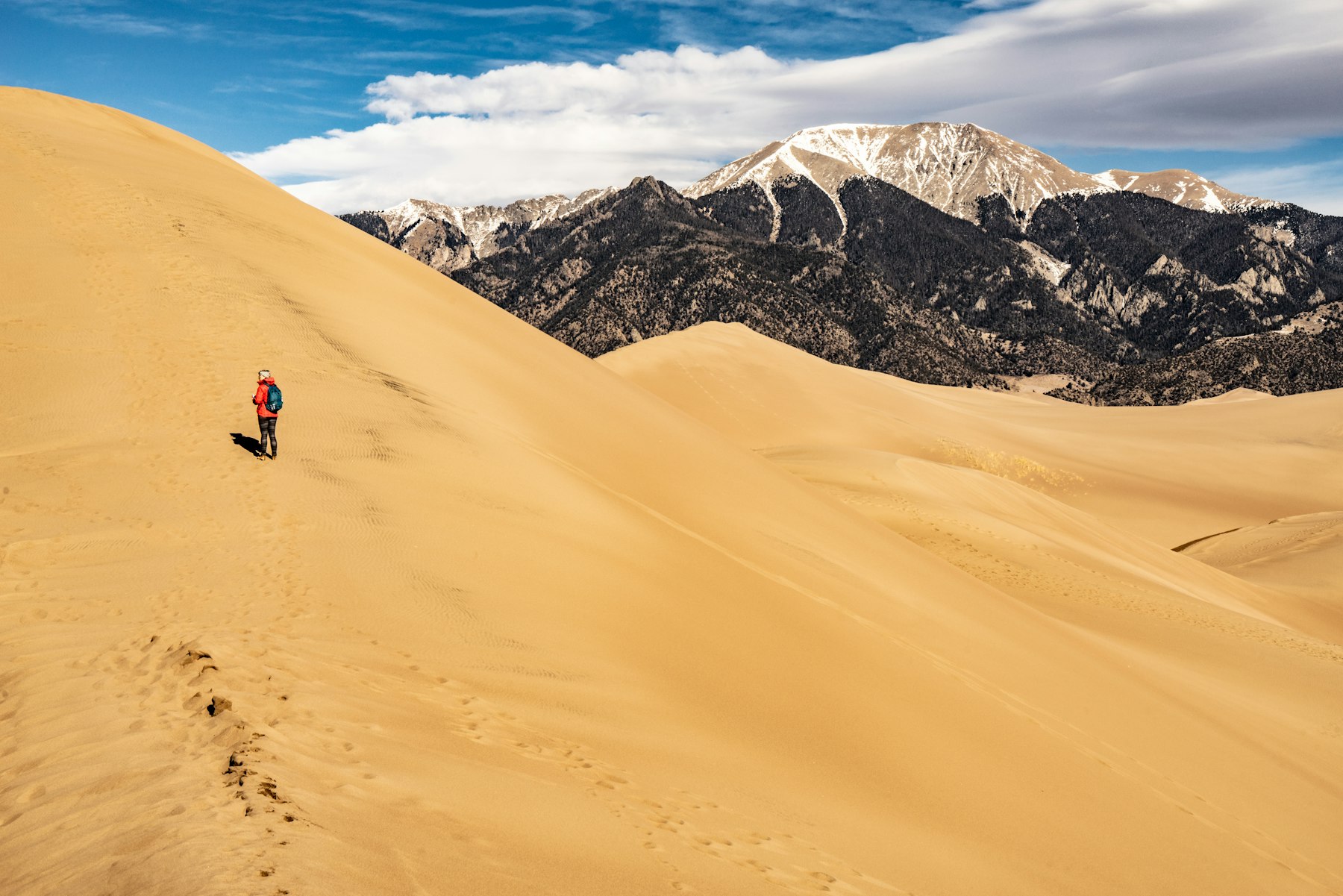 A person walking up a tall sand dune with a snow capped peak behind at Great Sand Dunes