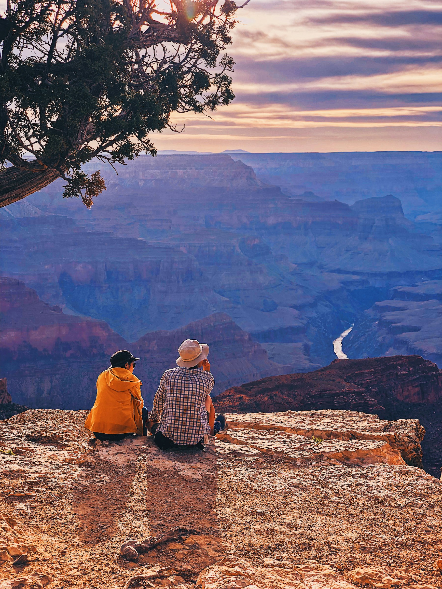 Two people sitting on the rim of the Grand Canyon looking down at the Colorado River