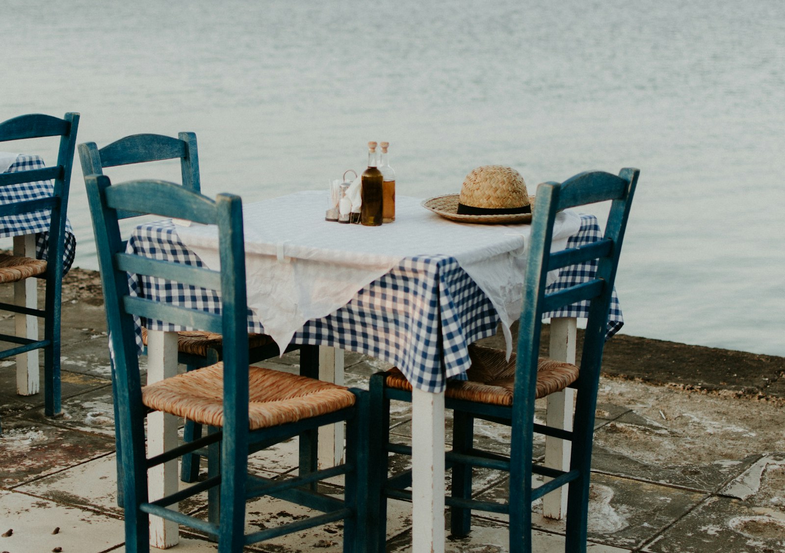 A taverna table with a checked cloth and blue wooden chairs set on a stone terrace beside the sea