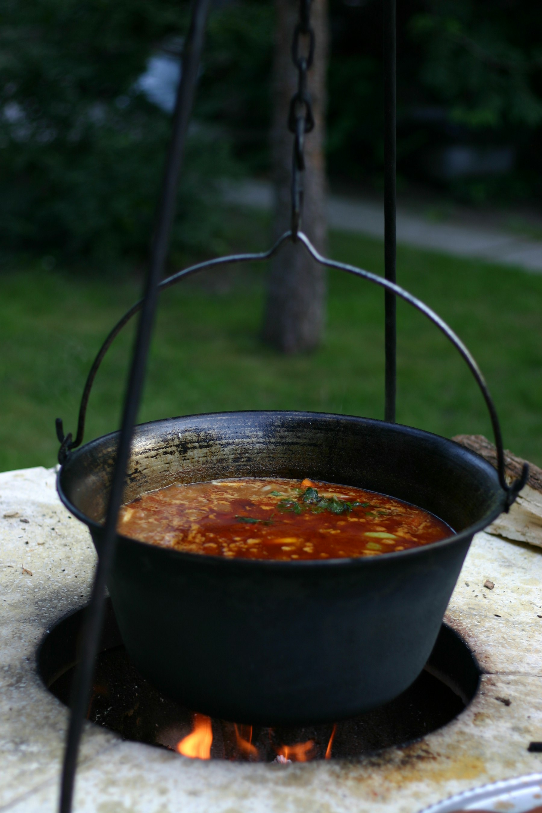 A black cast iron kettle hanging over an open fire filled with red paprika goulash soup