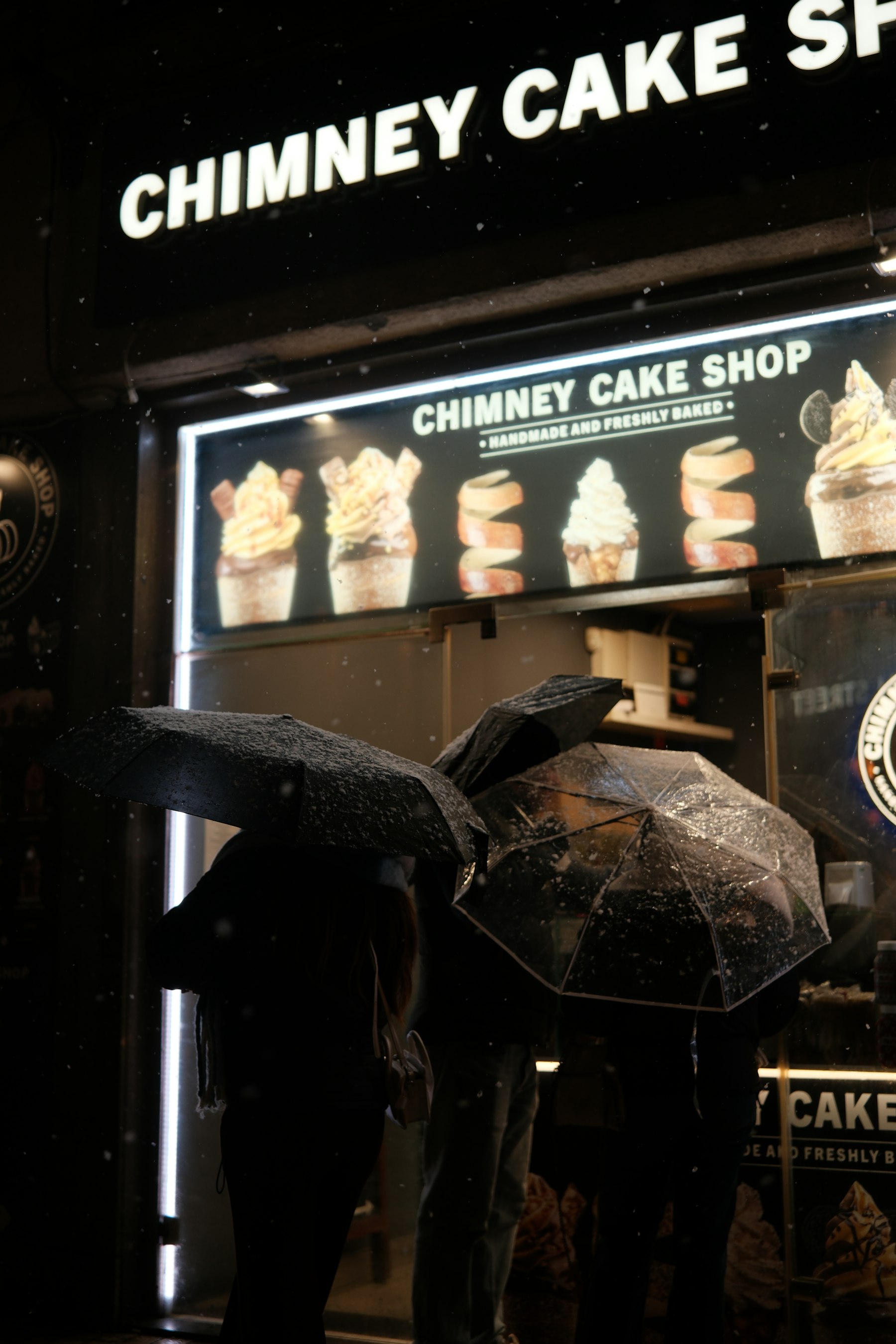 Chimney cakes turning on rollers under a lit sign reading Chimney Cake at a street stall