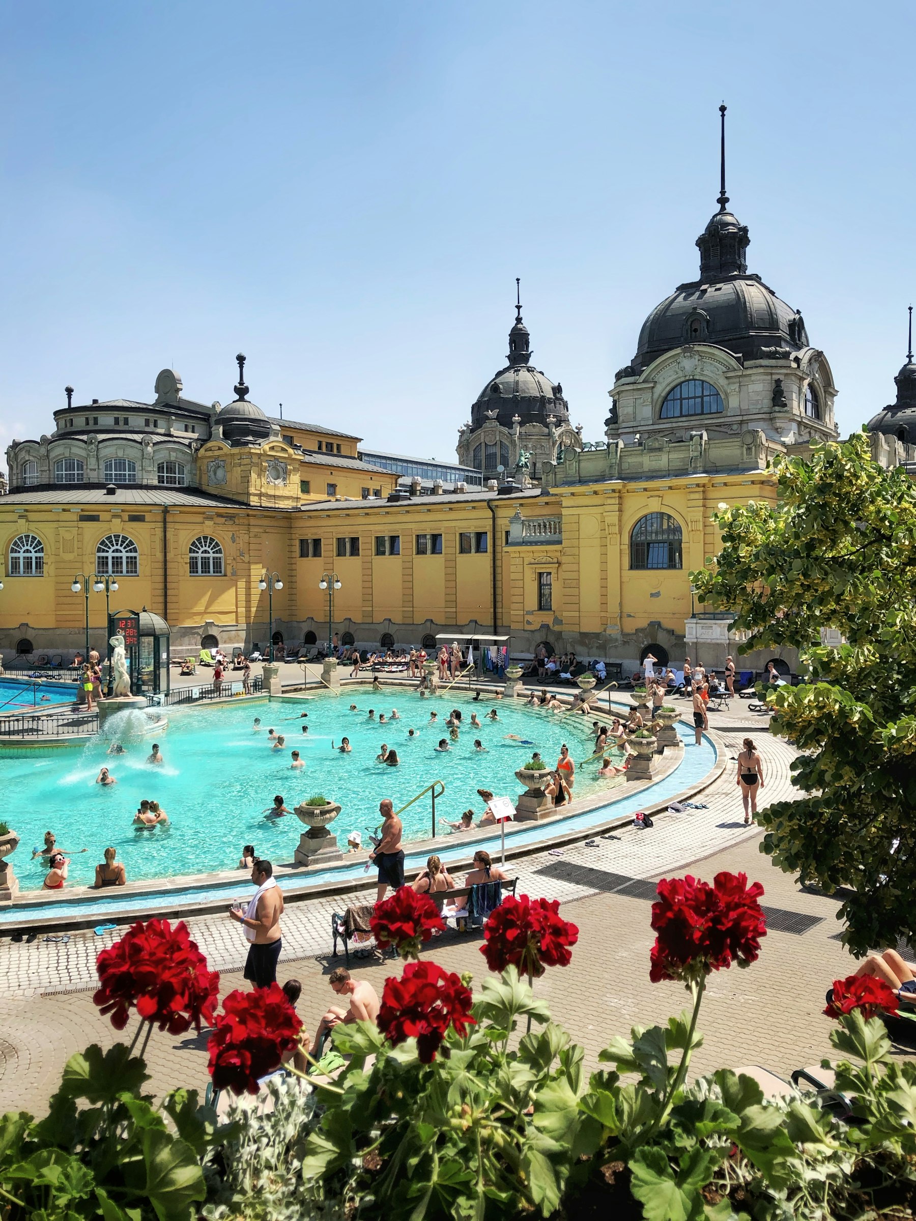 Bathers in the steaming turquoise outdoor pool of the Szechenyi thermal baths in Budapest below yellow neo baroque buildings