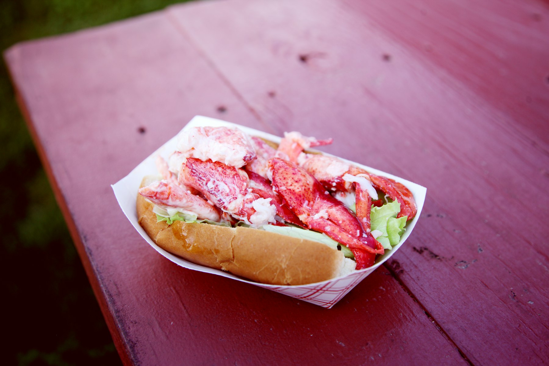 A lobster roll in a paper tray filled with chunks of lobster meat on a red picnic table