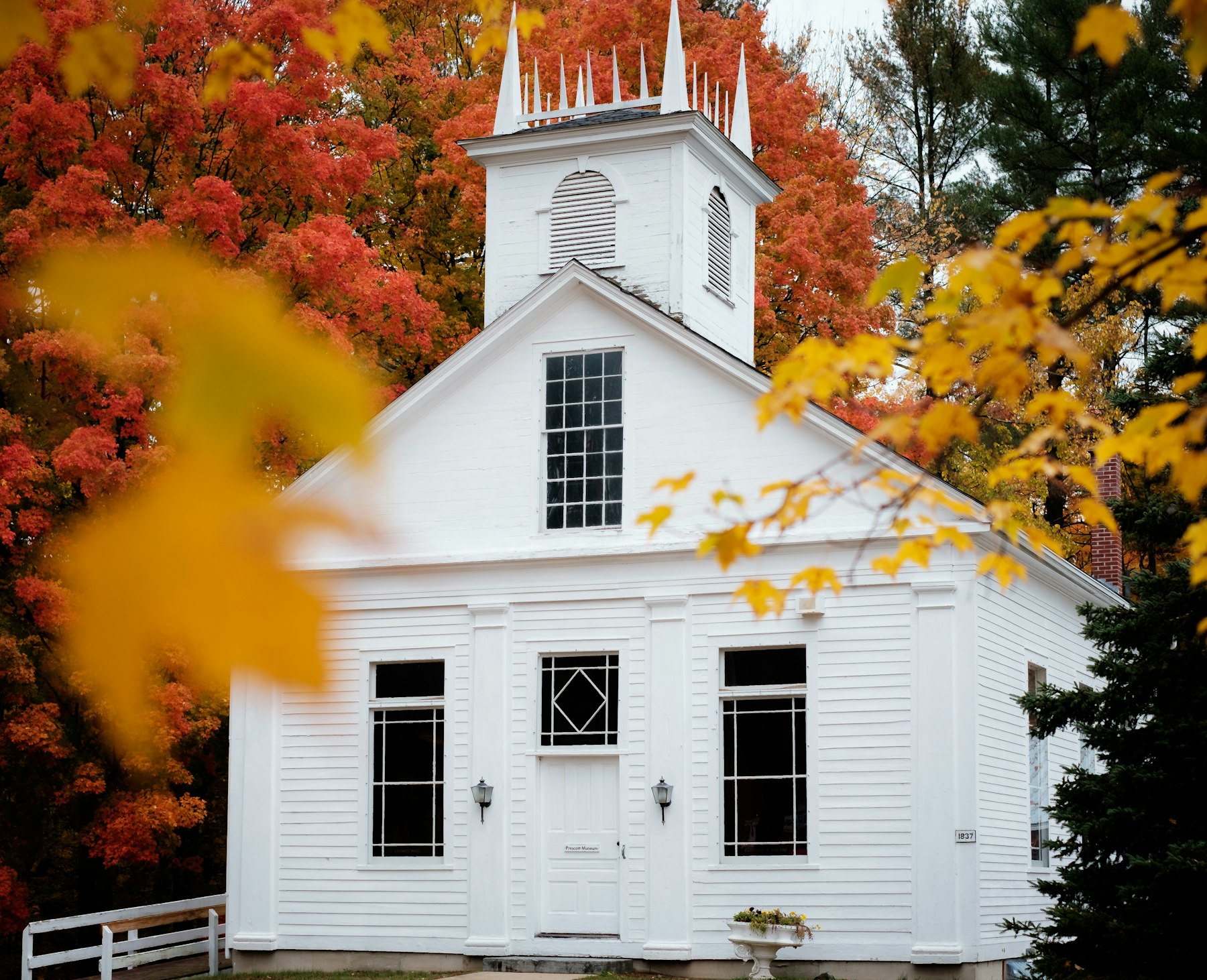 A white New England meeting house framed by orange and yellow autumn leaves