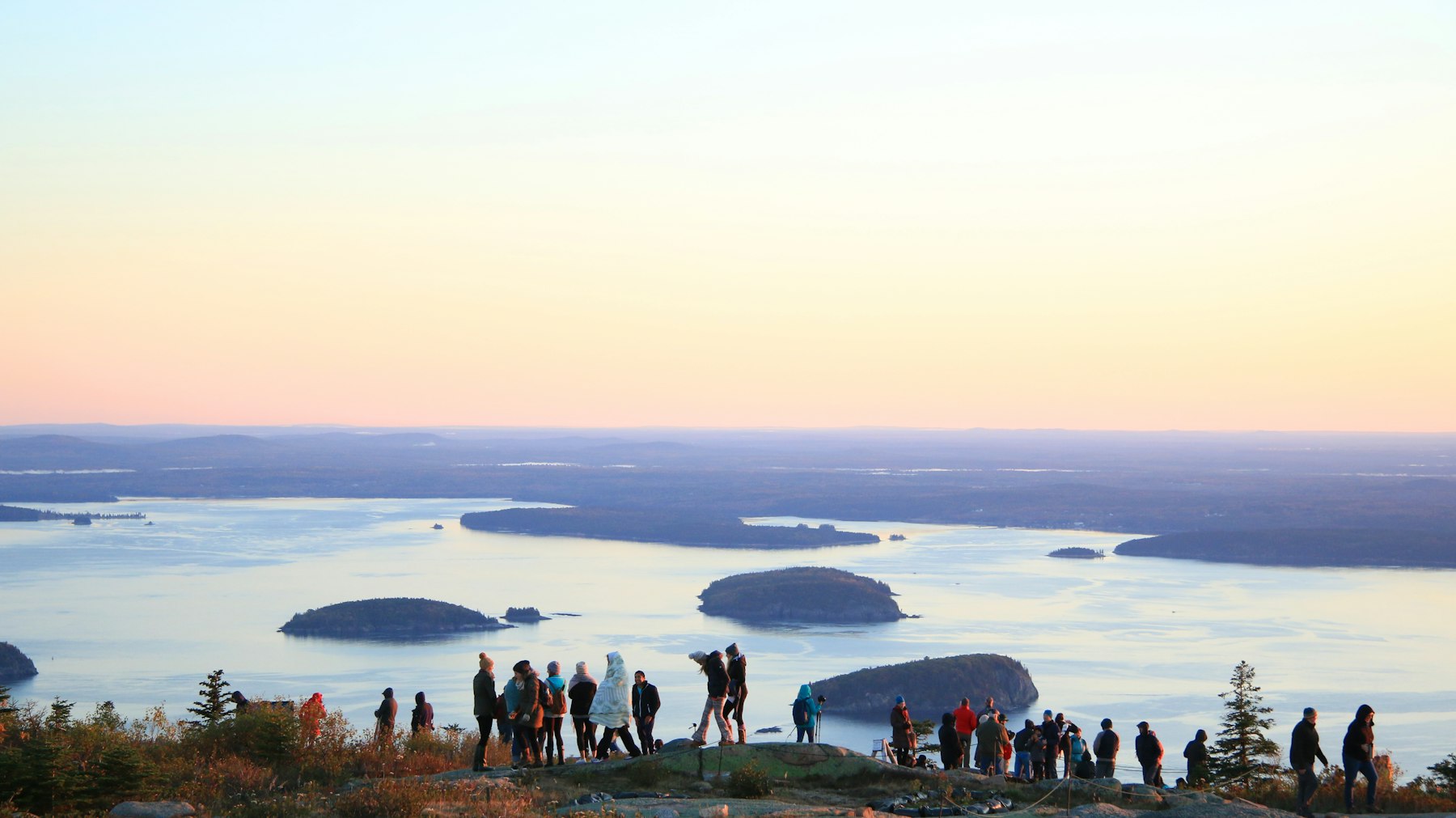 A crowd of people watching sunrise from the summit of Cadillac Mountain above islands in Acadia