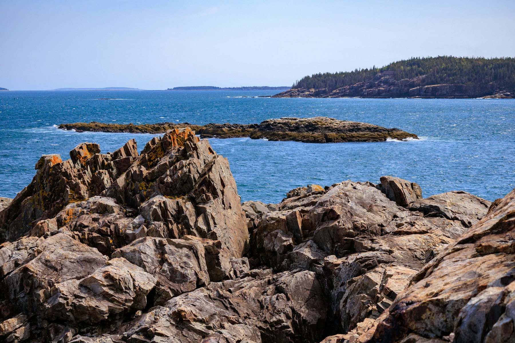 Rough pink granite ledges dropping into deep blue water with spruce covered islands beyond in Acadia