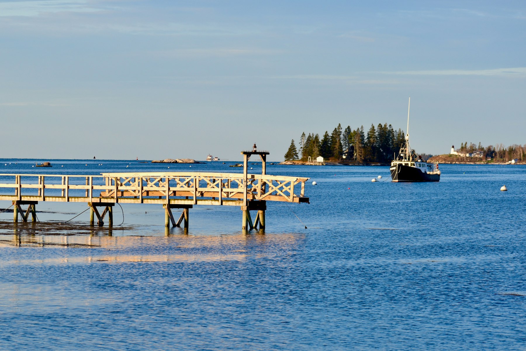 A wooden pier reaching into calm water with a boat and spruce islands at Boothbay Harbor Maine