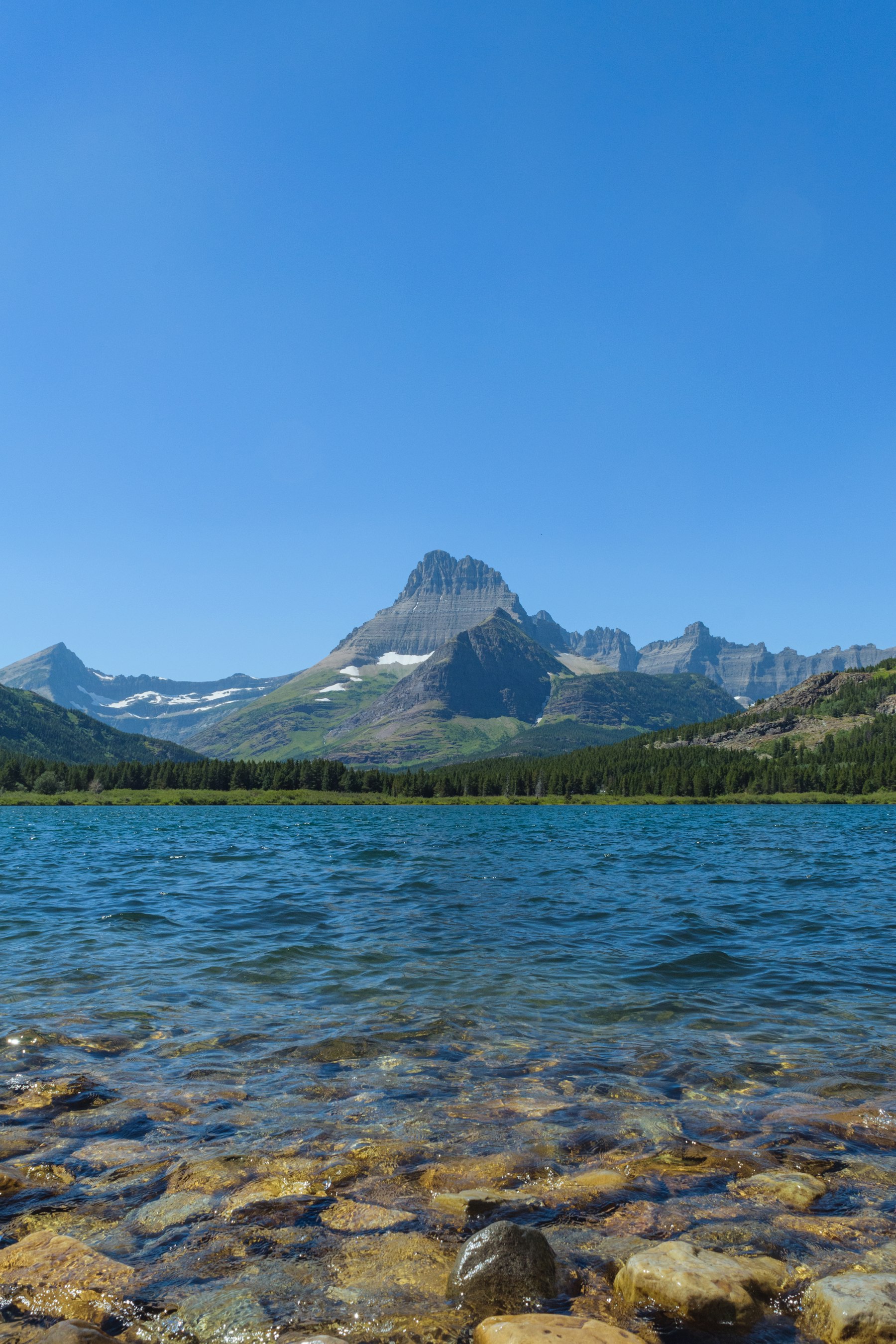 Coloured stones under clear lake water with a jagged peak behind in Glacier National Park Montana