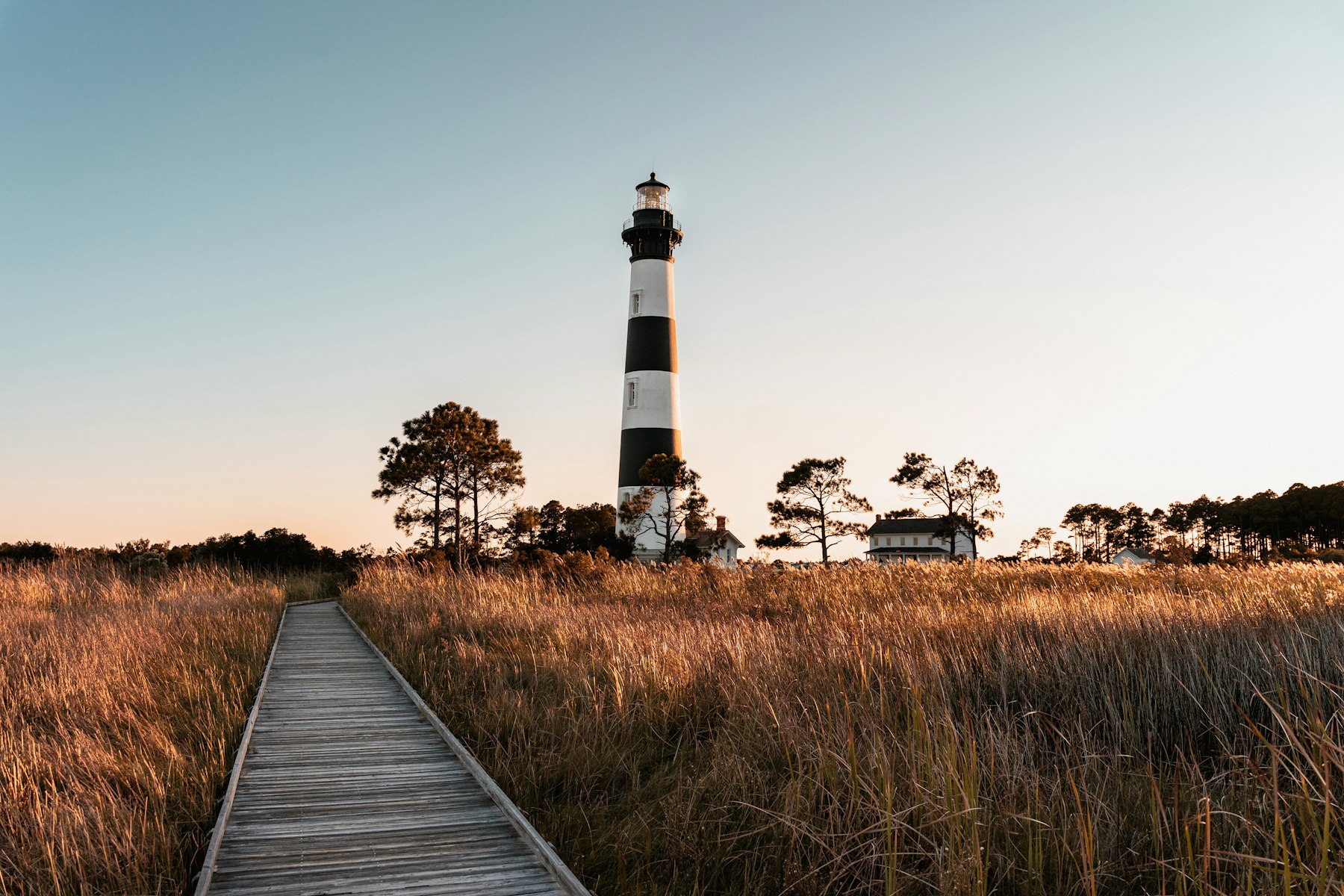 A wooden boardwalk crossing golden marsh grass toward the Bodie Island Lighthouse at sunset