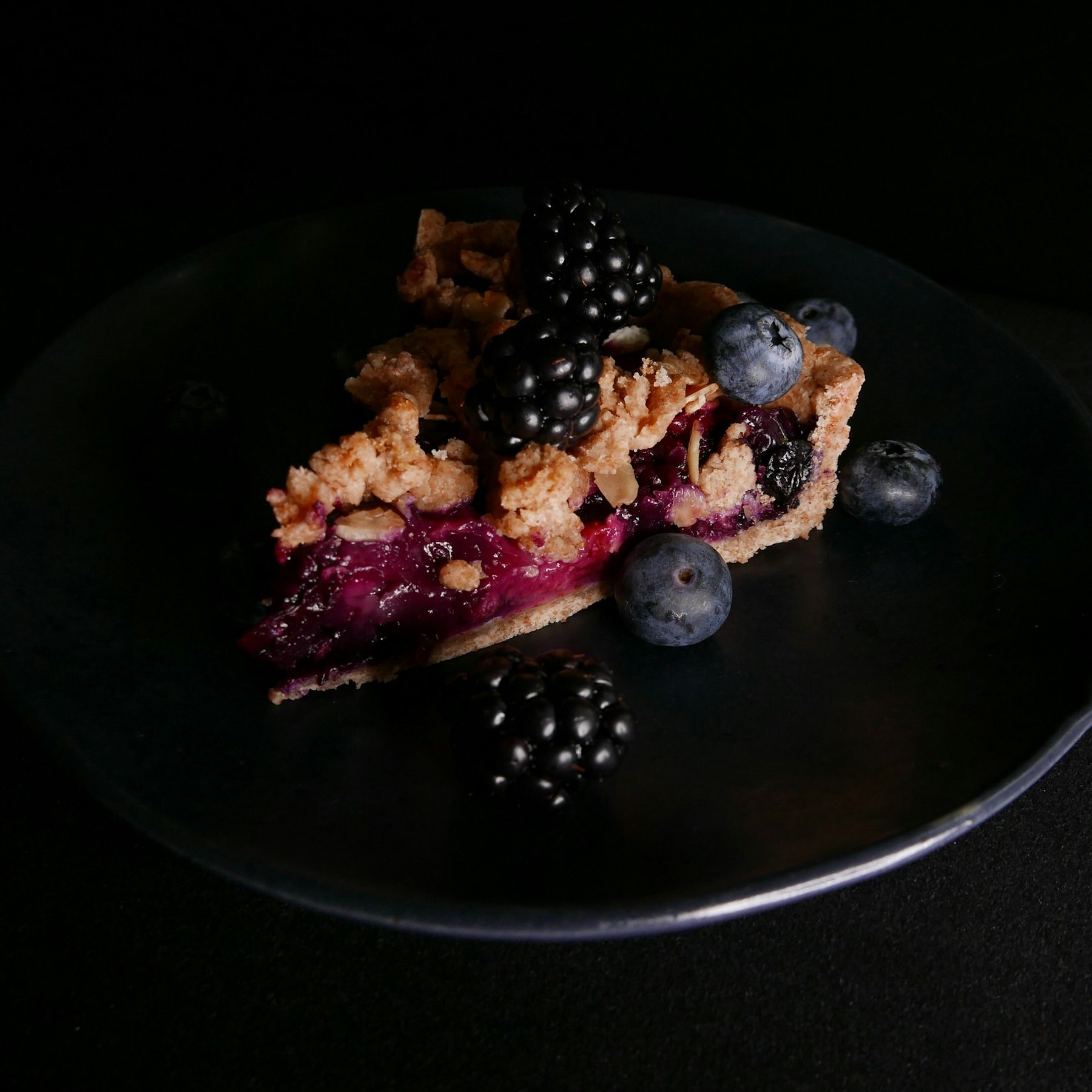A slice of blackberry pie with fresh berries on a dark plate
