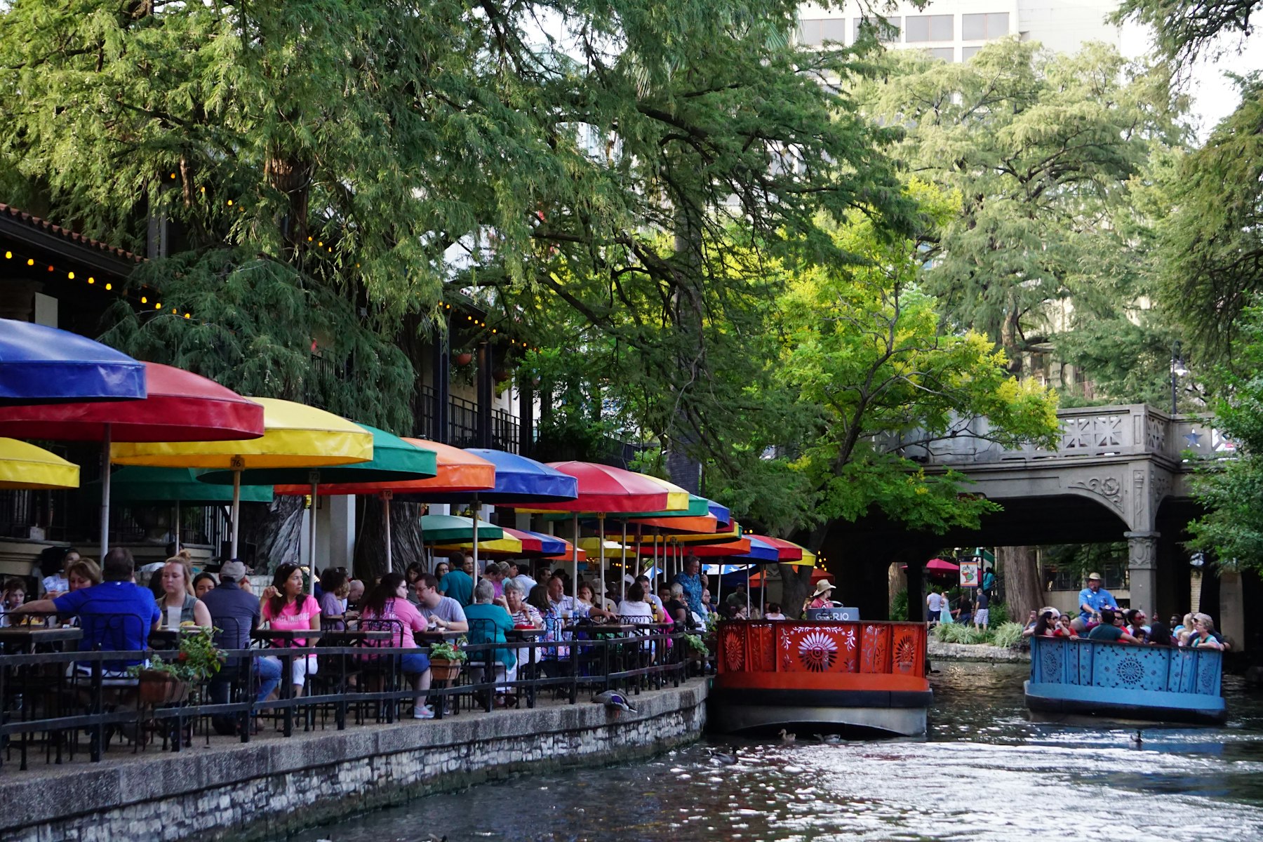 A red river barge carrying passengers past coloured umbrellas on the San Antonio River Walk