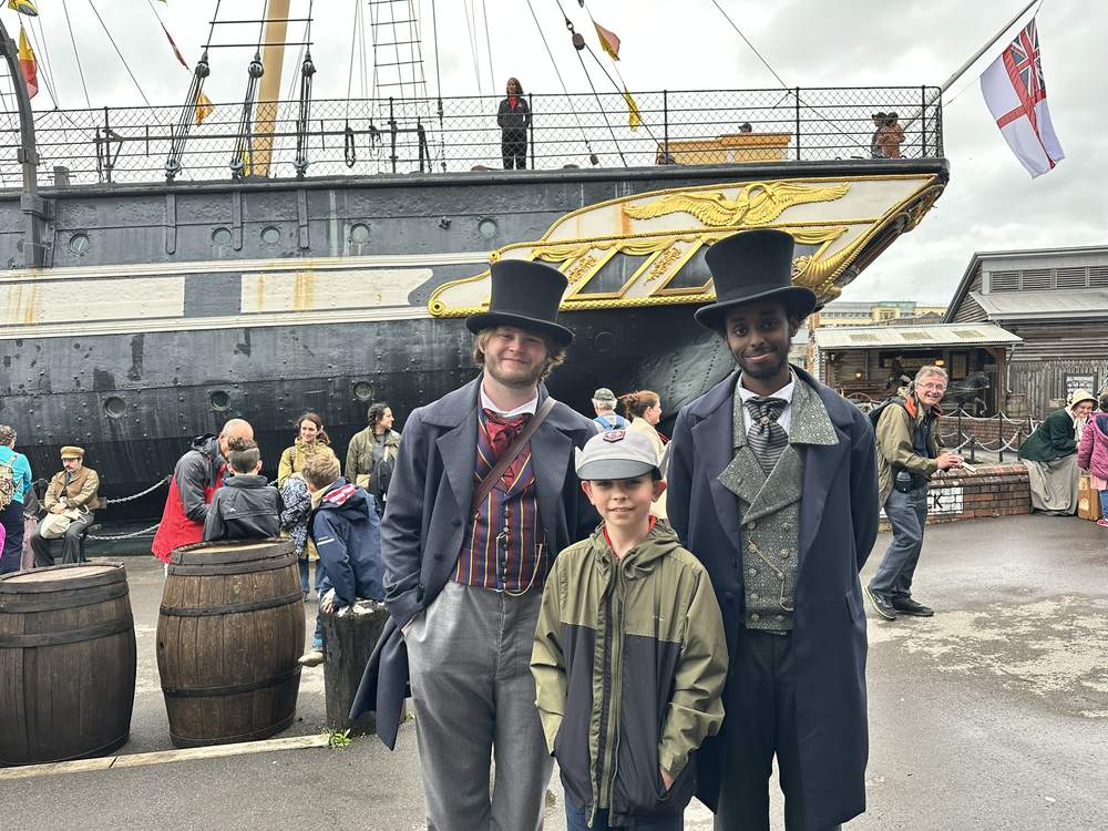 Two costumed Victorian guides in top hats standing in front of the black hull and gilded figurehead of the SS Great Britain in Bristol