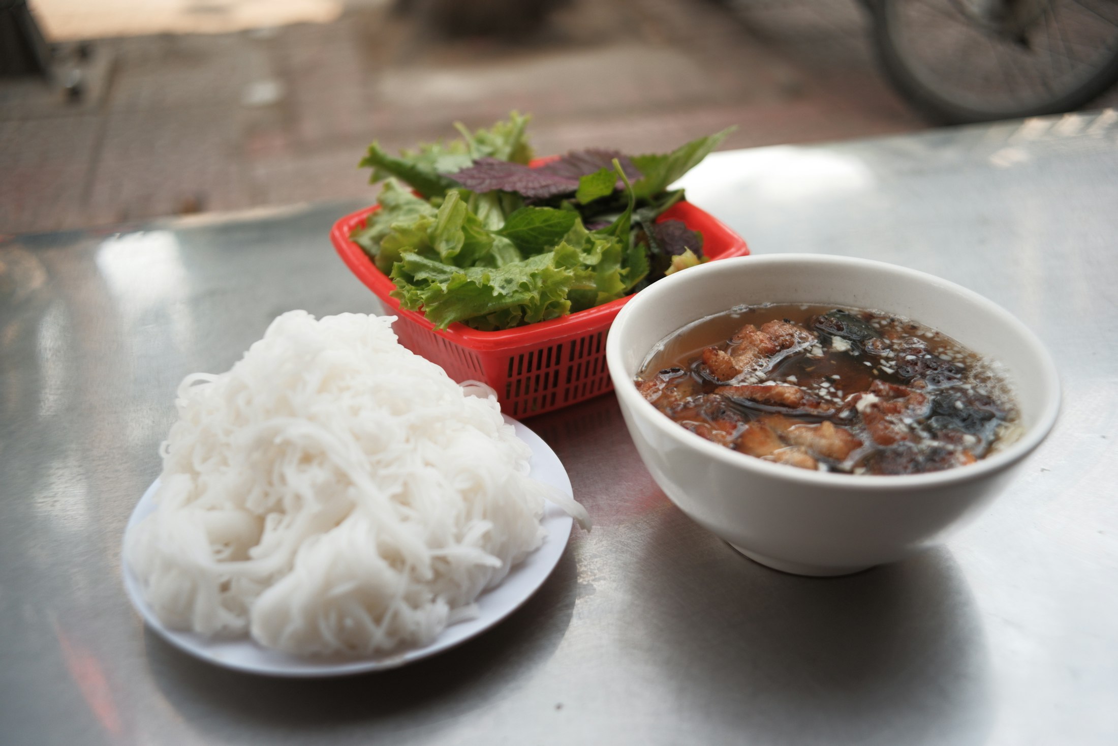 Bun cha, a plate of rice vermicelli beside a bowl of grilled pork in broth with a basket of fresh herbs