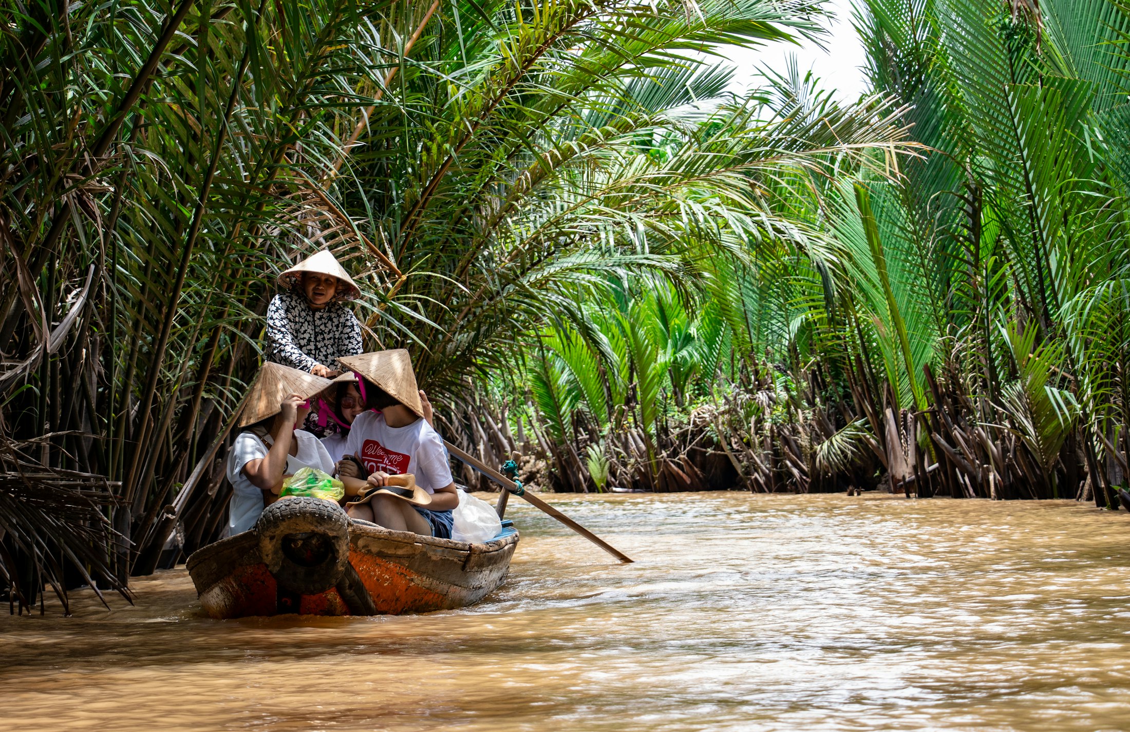 A woman in a conical hat rowing a narrow sampan along a palm lined channel in the Mekong Delta