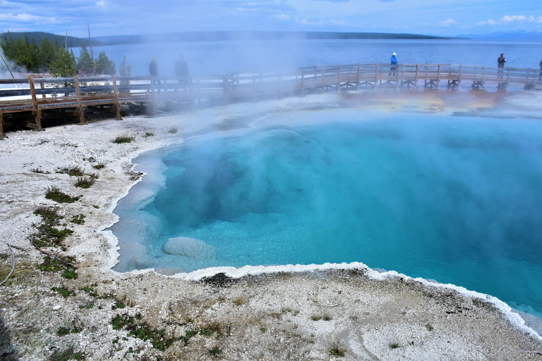 Visitors on the boardwalk at West Thumb Geyser Basin beside a deep blue pool on the shore of Yellowstone Lake